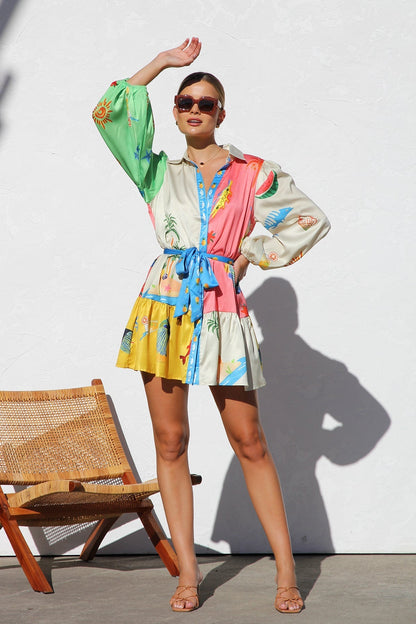 A woman with blonde hair in a bun smiles while wearing a colorful, long-sleeve mini shirt dress with a tropical print, belted at the waist, standing against a white textured wall.