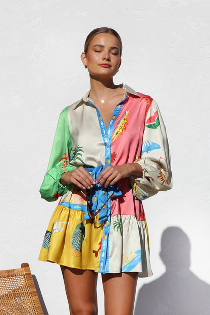 A woman with blonde hair in a bun smiles while wearing a colorful, long-sleeve mini shirt dress with a tropical print, belted at the waist, standing against a white textured wall.