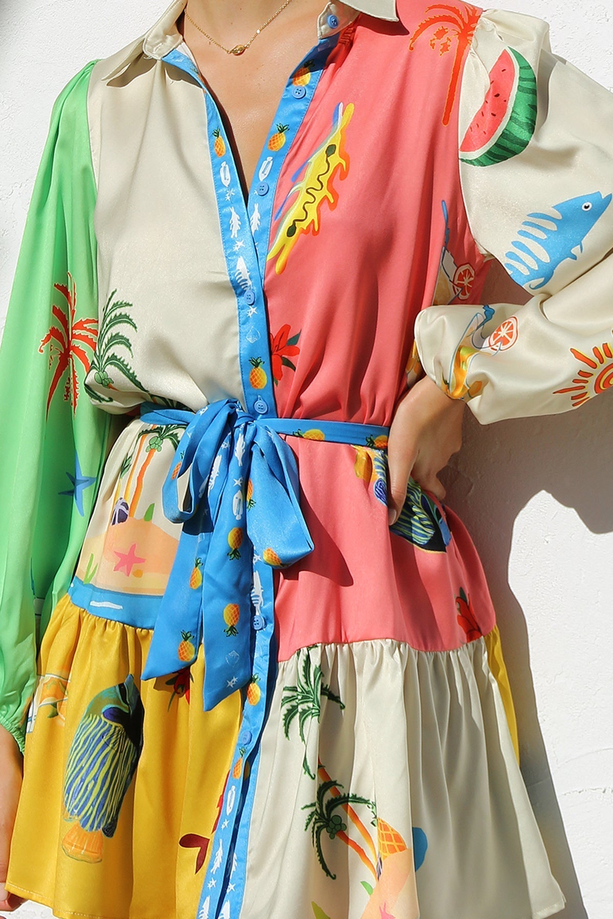 A woman with her hair in a sleek bun smiles while wearing a multi-patterned, long-sleeve tropical print mini shirt dress, standing against a white textured wall.