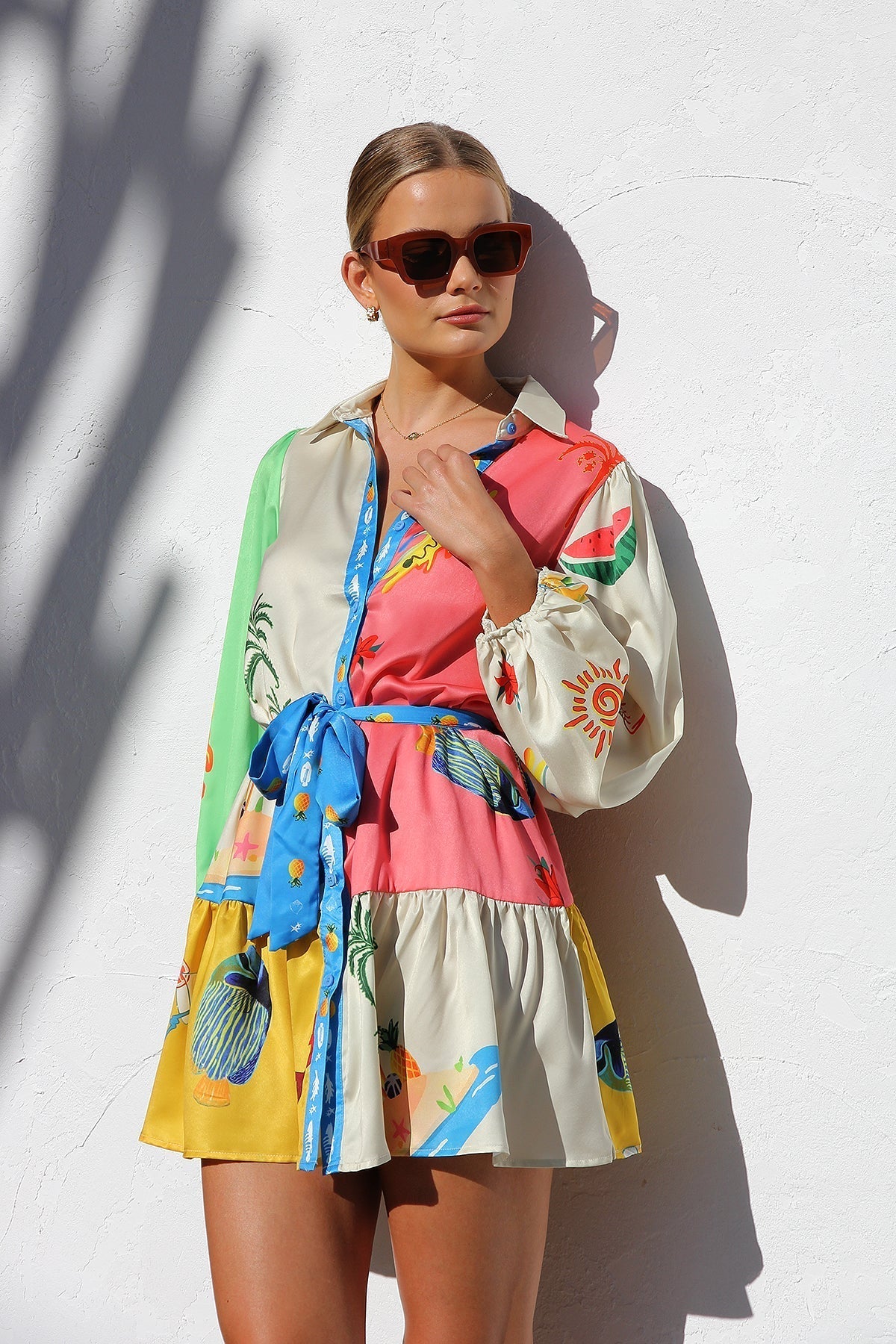 A woman with her hair in a sleek bun smiles while wearing a multi-patterned, long-sleeve tropical print mini shirt dress, standing against a white textured wall.
