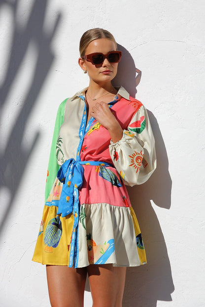 A woman with her hair in a sleek bun smiles while wearing a multi-patterned, long-sleeve tropical print mini shirt dress, standing against a white textured wall.