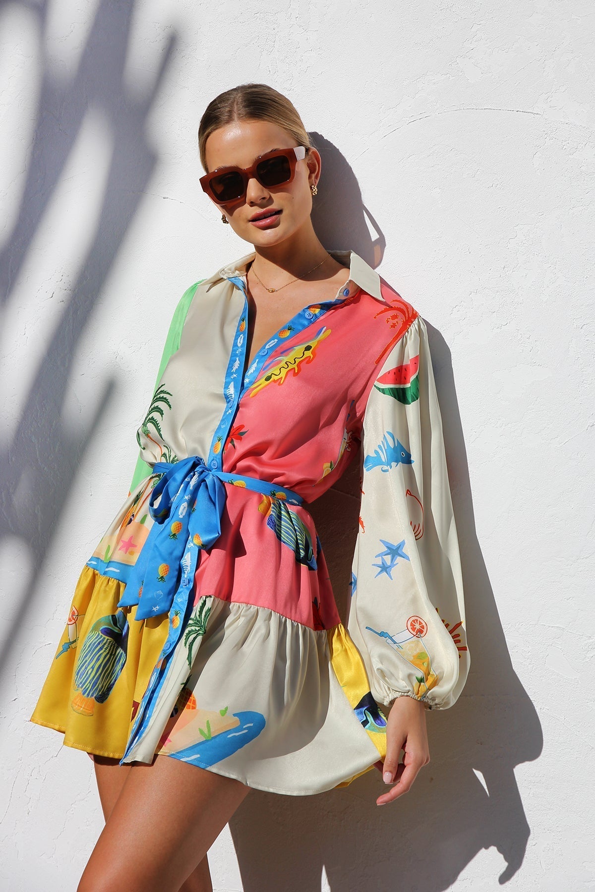 A woman with blonde hair in a bun smiles while wearing a colorful, long-sleeve mini shirt dress with a tropical print, belted at the waist, standing against a white textured wall.