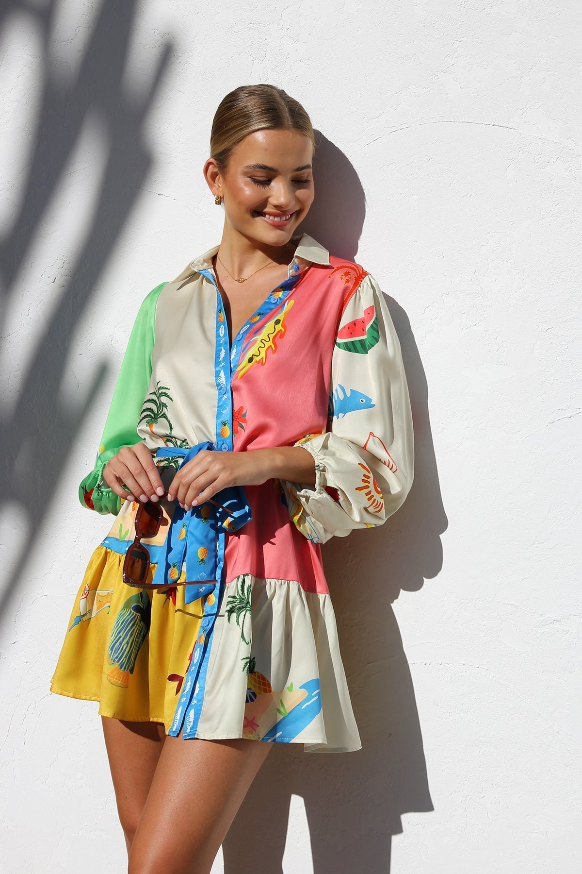 A woman with blonde hair in a bun smiles while wearing a colorful, long-sleeve mini shirt dress with a tropical print, belted at the waist, standing against a white textured wall.