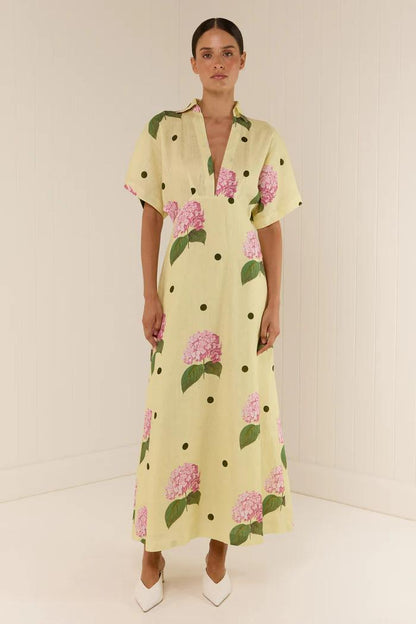 Full-length view of a model wearing a yellow collared maxi dress with short sleeves, a deep V-neck, and a pattern of pink hydrangeas and black polka dots.