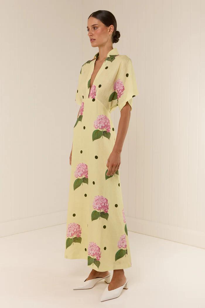 Full-length view of a model wearing a yellow collared maxi dress with short sleeves, a deep V-neck, and a pattern of pink hydrangeas and black polka dots.