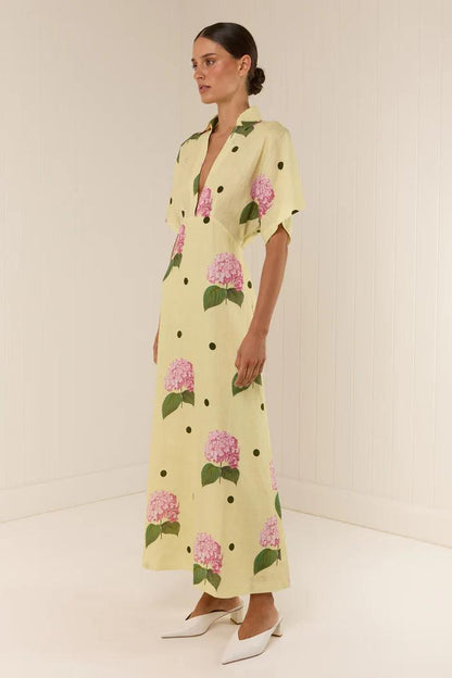 Full-length view of a model wearing a yellow collared maxi dress with short sleeves, a deep V-neck, and a pattern of pink hydrangeas and black polka dots.
