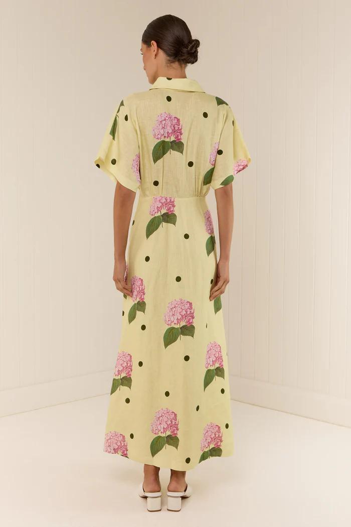 Full-length view of a model wearing a yellow collared maxi dress with short sleeves, a deep V-neck, and a pattern of pink hydrangeas and black polka dots.
