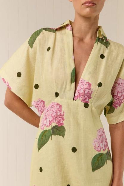 Full-length view of a model wearing a yellow collared maxi dress with short sleeves, a deep V-neck, and a pattern of pink hydrangeas and black polka dots.