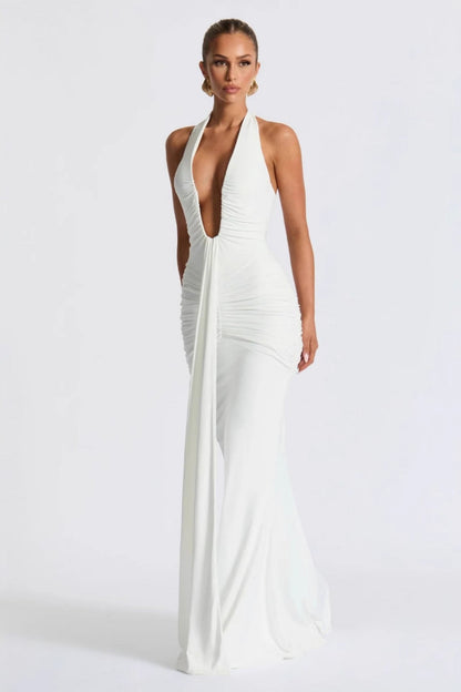 White - A woman models a form-fitting, deep red halterneck maxi dress with a plunging neckline and extensive ruching, standing elegantly in a formal garden setting.