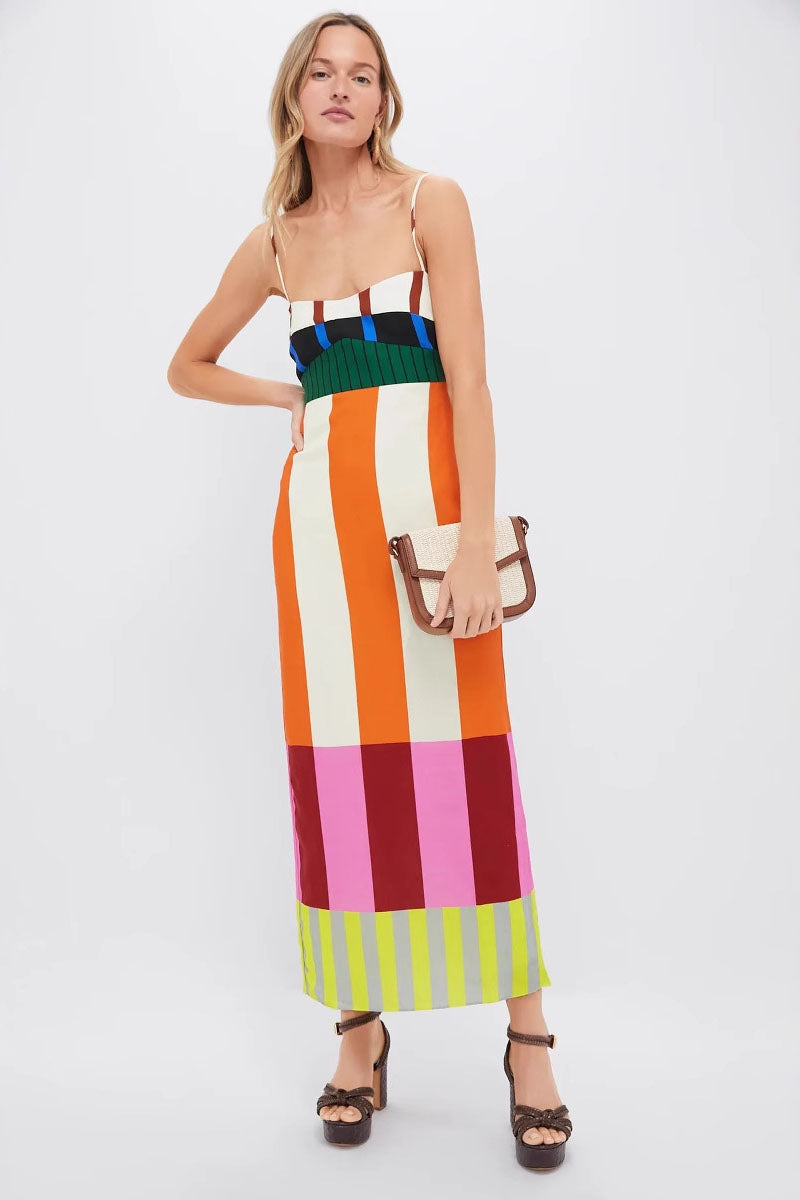 A full-length shot of a woman modeling a sleeveless midi dress with spaghetti straps. The dress has a straight, column silhouette and features a vibrant, multi-colored pattern of vertical stripes and color-blocked panels at the bodice and hem.