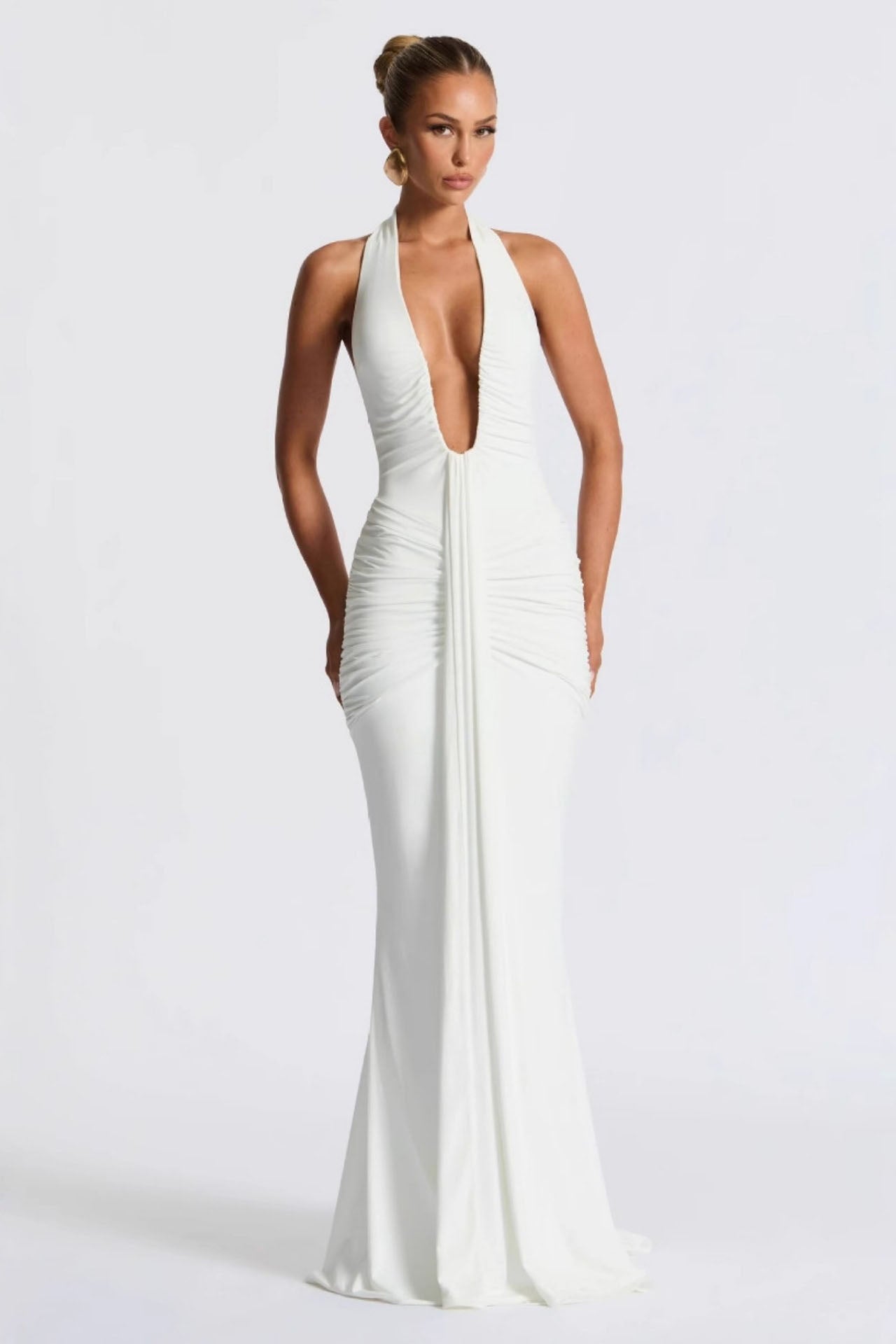 White - A woman models a form-fitting, deep red halterneck maxi dress with a plunging neckline and extensive ruching, standing elegantly in a formal garden setting.