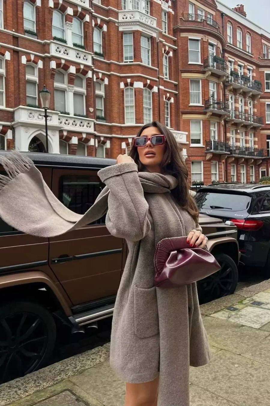 Khaki - A woman wearing a thick, taupe oversized longline winter coat featuring a built-in scarf neckline, dark button closures, and large patch pockets.