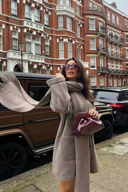 Khaki - A woman wearing a thick, taupe oversized longline winter coat featuring a built-in scarf neckline, dark button closures, and large patch pockets.