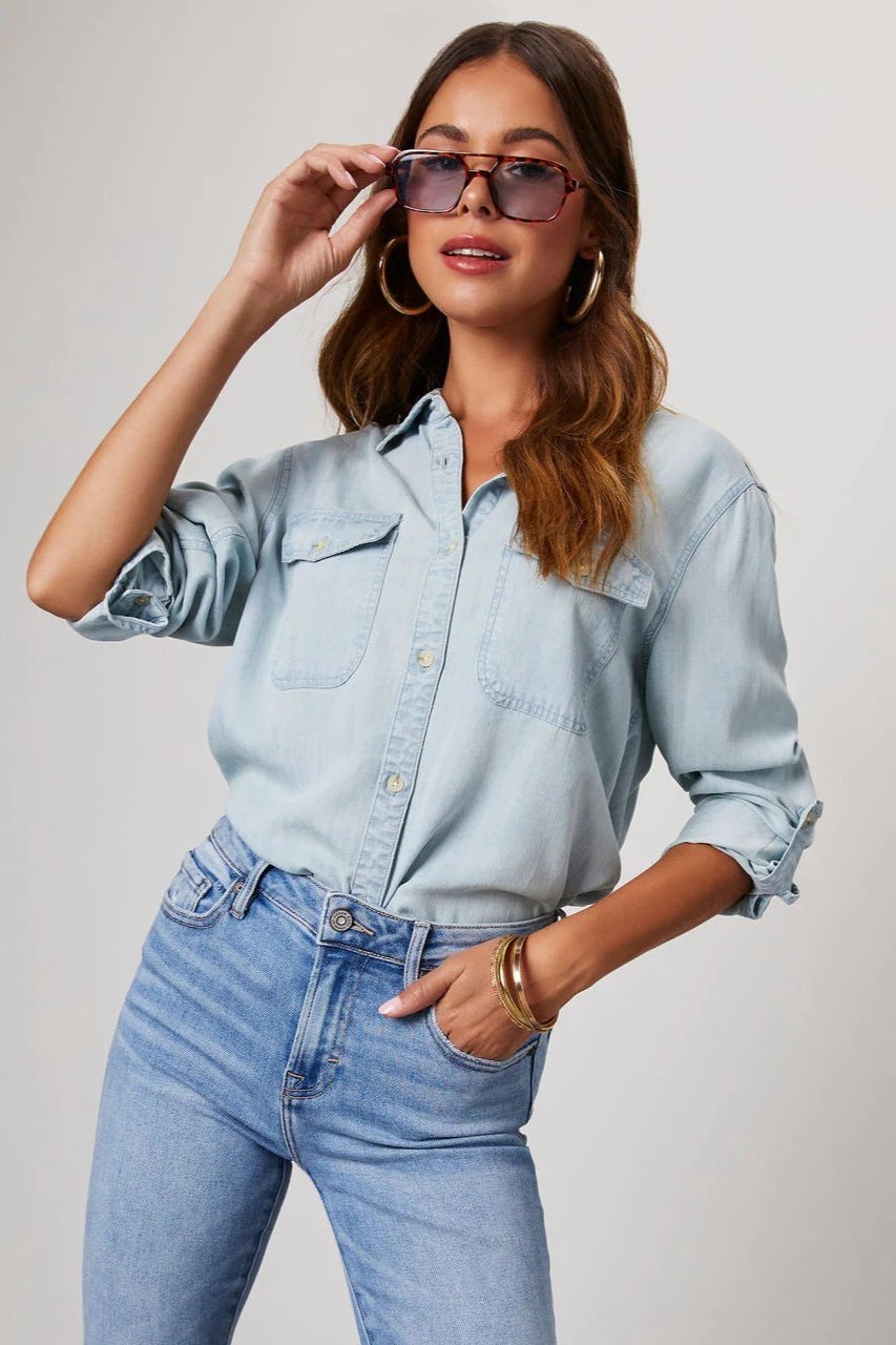 Blue - Women's light blue denim button-down shirt featuring a point collar, dual chest flap pockets, and rolled sleeves secured with button tabs, styled casually.