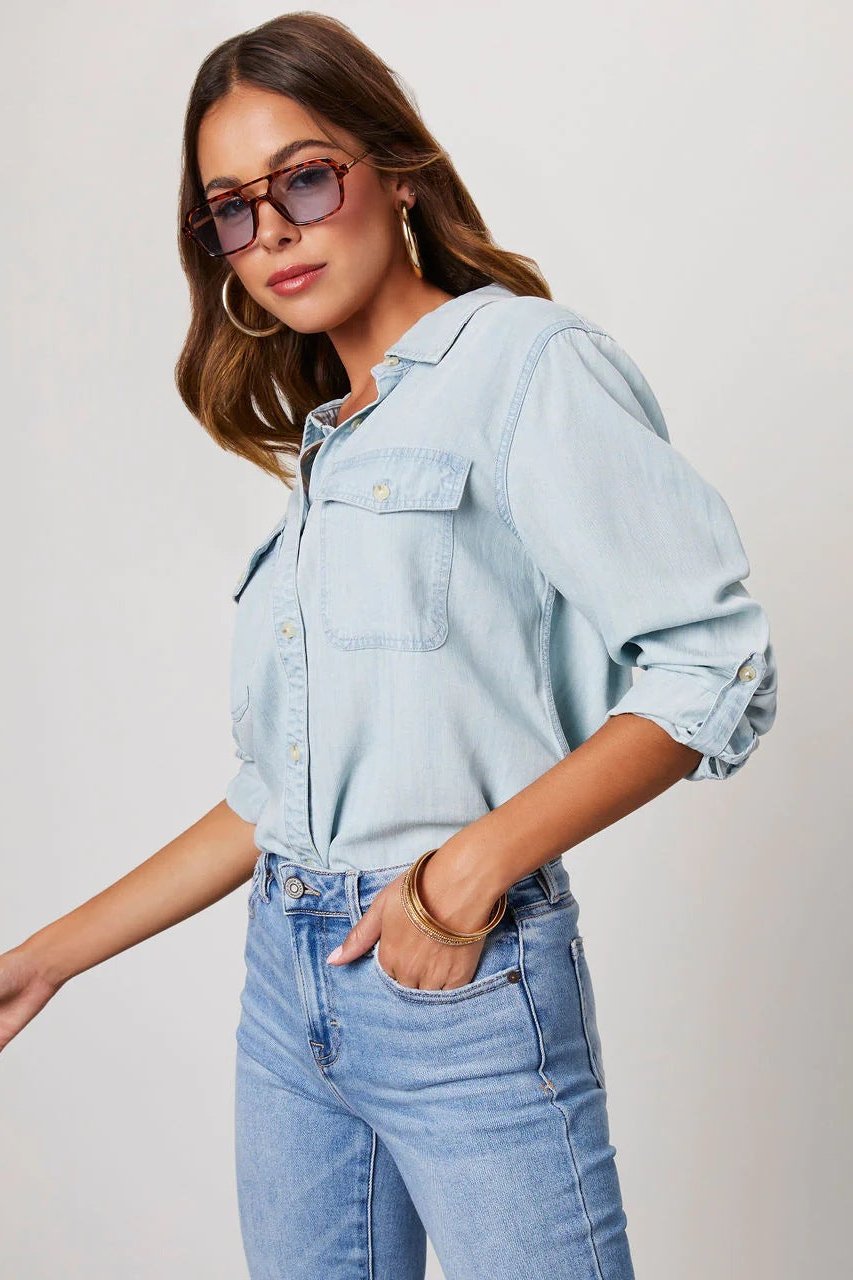 Light Blue - Women's light blue denim button-down shirt featuring a point collar, dual chest flap pockets, and rolled sleeves secured with button tabs, styled casually.