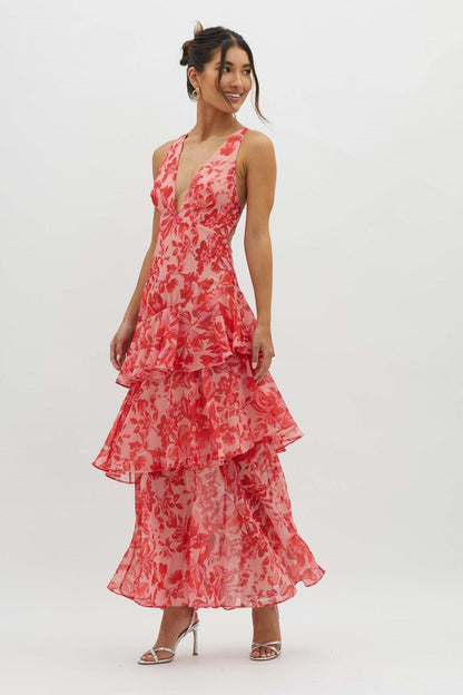 A woman models a sleeveless maxi dress with a deep V-neck and a tiered, ruffled skirt, featuring a vibrant red and pink floral print on a light pink background.