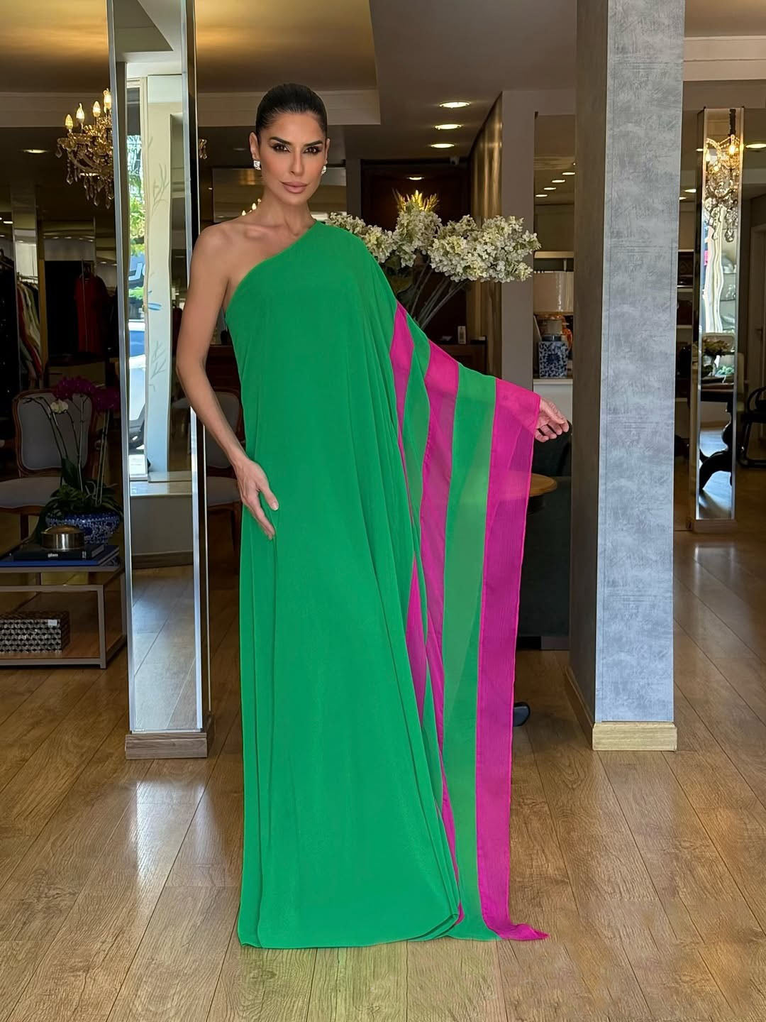 A woman models a vibrant green, one-shoulder, floor-length gown against a white background. The gown has a dramatic, flowing sleeve with a bright pink contrasting interior panel.