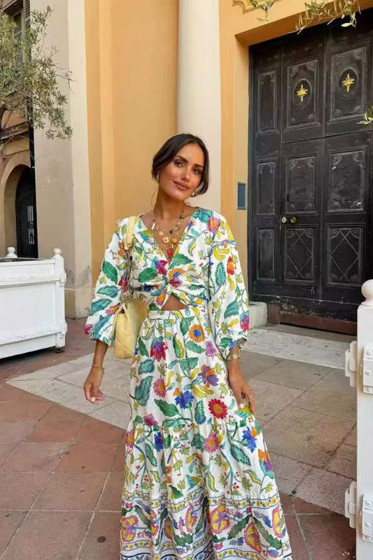 A woman with dark hair in an updo stands on a stone patio, wearing a long-sleeve, white maxi dress with a vibrant multi-color floral print, a v-neck, a twist-front bodice, and a waist cutout.