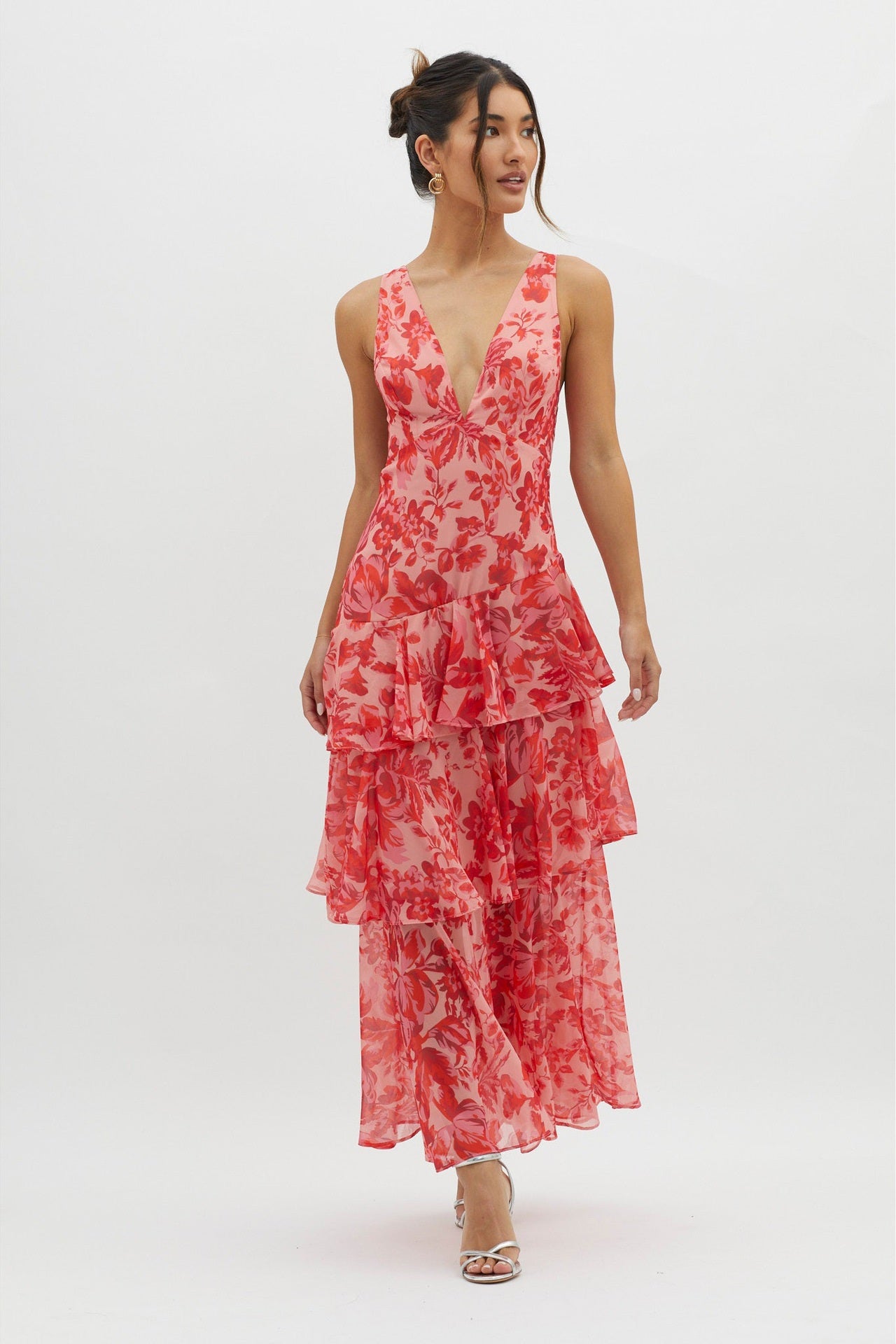 A woman models a sleeveless maxi dress with a deep V-neck and a tiered, ruffled skirt, featuring a vibrant red and pink floral print on a light pink background.