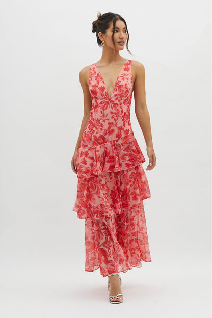 A woman models a sleeveless maxi dress with a deep V-neck and a tiered, ruffled skirt, featuring a vibrant red and pink floral print on a light pink background.