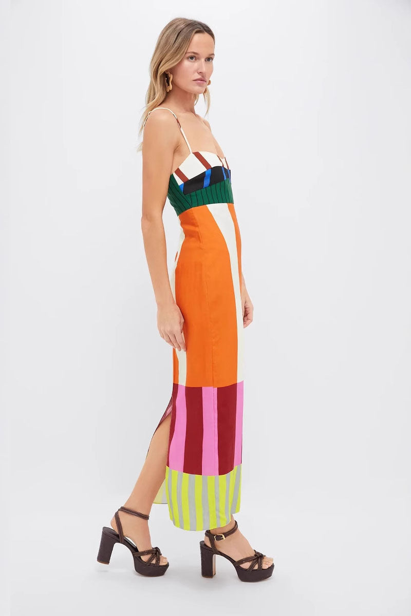 A full-length shot of a woman modeling a sleeveless midi dress with spaghetti straps. The dress has a straight, column silhouette and features a vibrant, multi-colored pattern of vertical stripes and color-blocked panels at the bodice and hem.