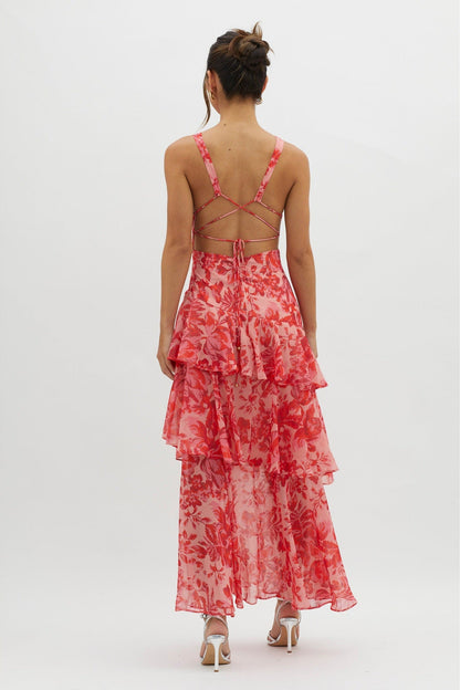 A woman models a sleeveless maxi dress with a deep V-neck and a tiered, ruffled skirt, featuring a vibrant red and pink floral print on a light pink background.