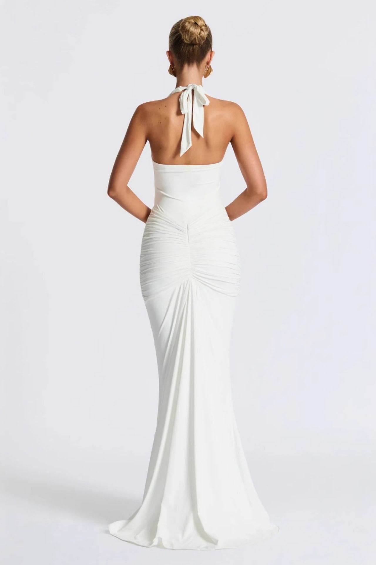 White - A woman models a form-fitting, deep red halterneck maxi dress with a plunging neckline and extensive ruching, standing elegantly in a formal garden setting.
