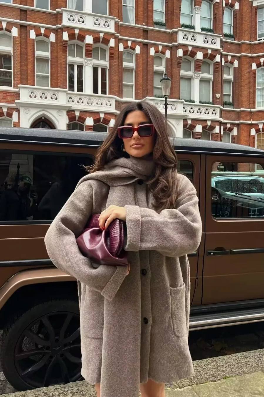 Khaki - A woman wearing a thick, taupe oversized longline winter coat featuring a built-in scarf neckline, dark button closures, and large patch pockets.