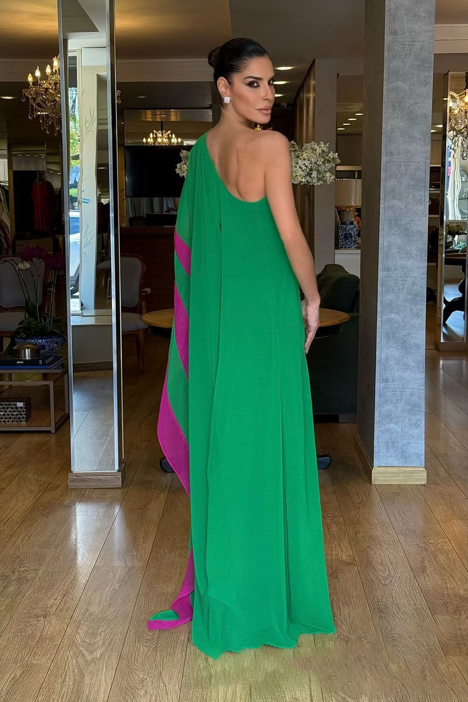 A woman models a vibrant green, one-shoulder, floor-length gown against a white background. The gown has a dramatic, flowing sleeve with a bright pink contrasting interior panel.