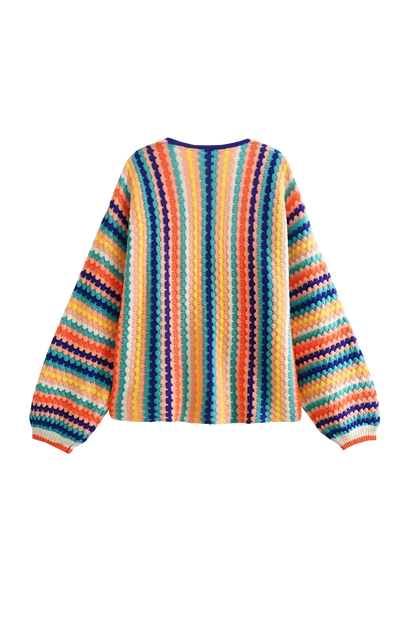 Rainbow - A woman wearing a vibrant, striped open-front crochet cardigan with ribbed cuffs, styled over a ribbed top and dark denim jeans on a city street.