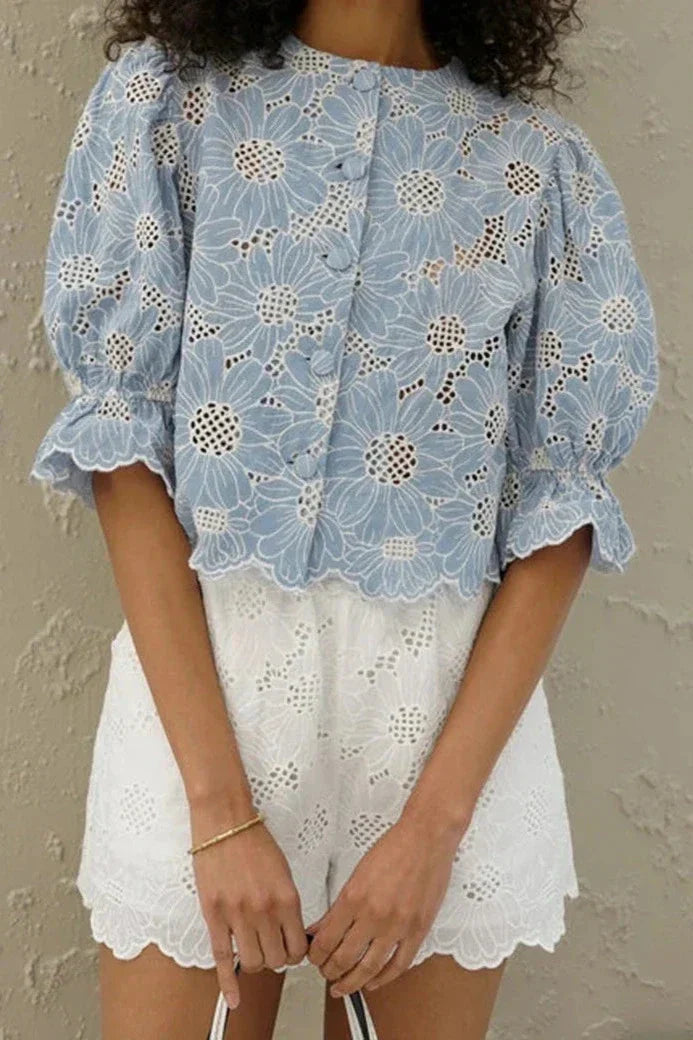 Woman in light blue floral eyelet blouse and white shorts holding straw bag.
