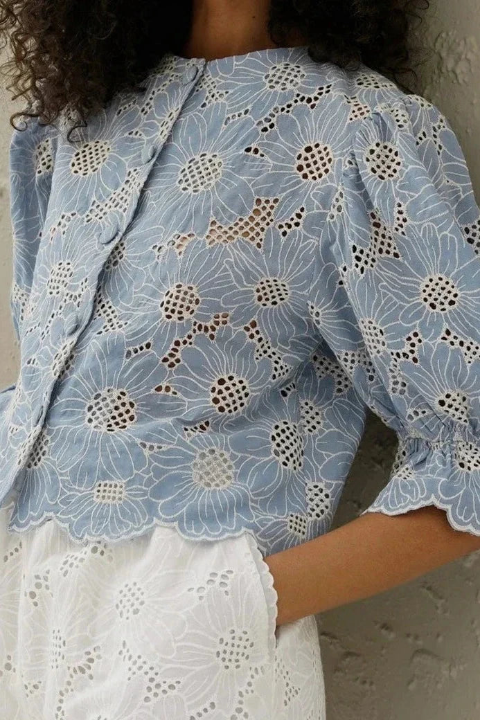 Woman in light blue floral eyelet blouse and white shorts holding straw bag.