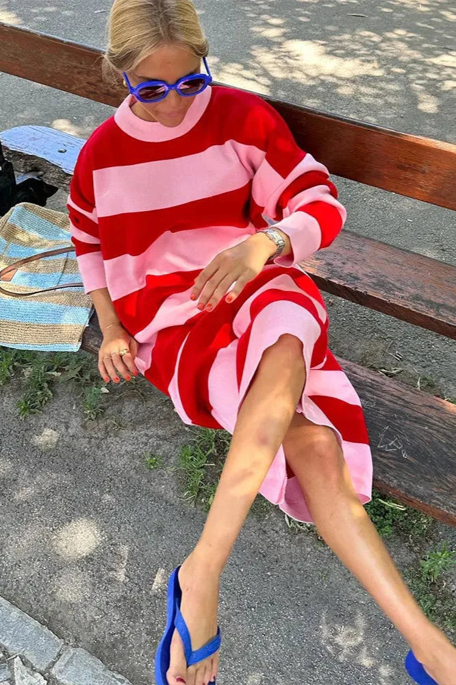 A woman standing on a sand dune, wearing a long-sleeved knit dress with bold red and pink horizontal stripes against a clear blue sky.