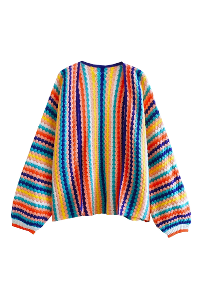 Rainbow - A woman wearing a vibrant, striped open-front crochet cardigan with ribbed cuffs, styled over a ribbed top and dark denim jeans on a city street.
