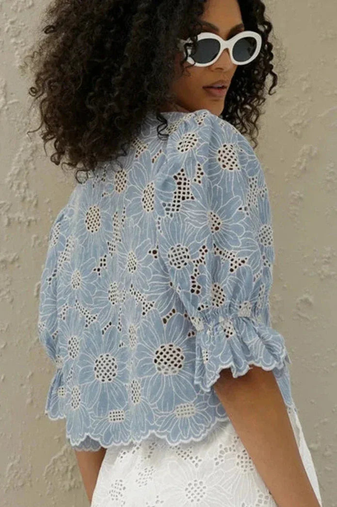 Woman in light blue floral eyelet blouse and white shorts holding straw bag.