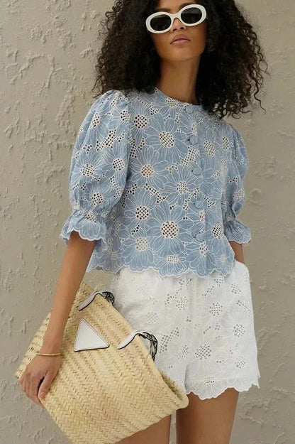 Woman in light blue floral eyelet blouse and white shorts holding straw bag.