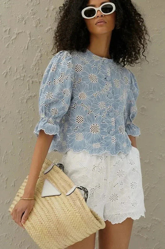 Woman in light blue floral eyelet blouse and white shorts holding straw bag.