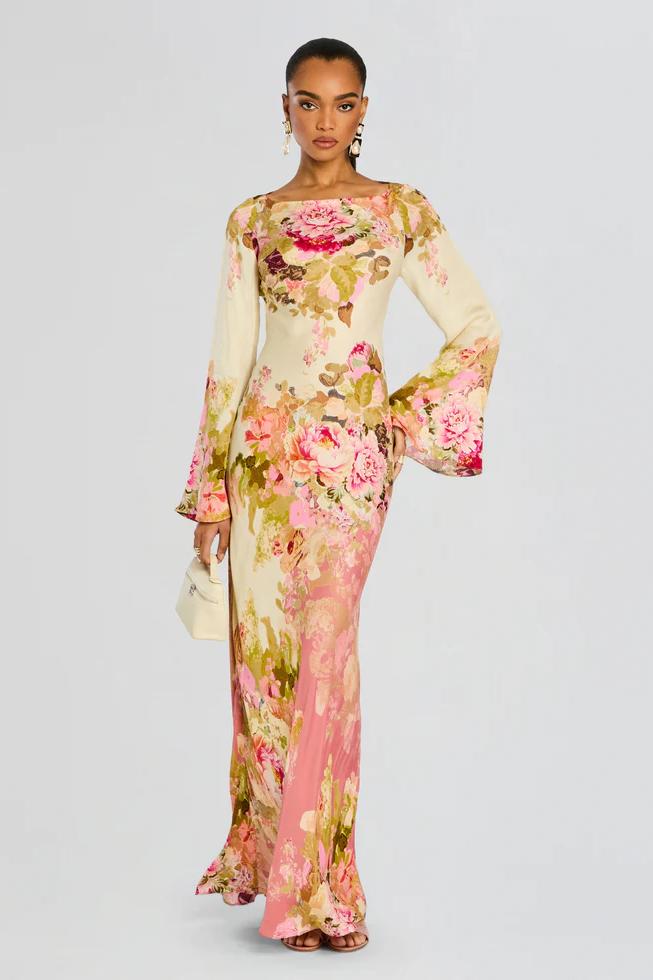 A model stands wearing a long-sleeved, floor-length column dress with a bateau neckline and dramatic flared bell sleeves. The cream-colored gown is adorned with a large-scale painterly floral print in shades of pink, rose, and olive green.