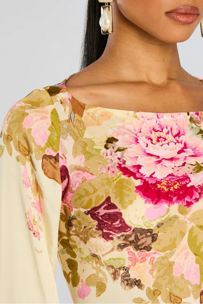 A model stands wearing a long-sleeved, floor-length column dress with a bateau neckline and dramatic flared bell sleeves. The cream-colored gown is adorned with a large-scale painterly floral print in shades of pink, rose, and olive green.