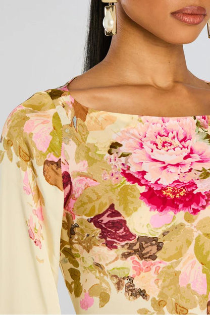 A model stands wearing a long-sleeved, floor-length column dress with a bateau neckline and dramatic flared bell sleeves. The cream-colored gown is adorned with a large-scale painterly floral print in shades of pink, rose, and olive green.