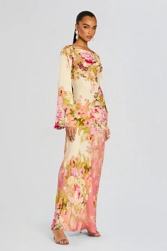 A model stands wearing a long-sleeved, floor-length column dress with a bateau neckline and dramatic flared bell sleeves. The cream-colored gown is adorned with a large-scale painterly floral print in shades of pink, rose, and olive green.