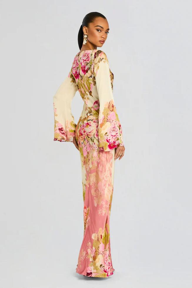 A model stands wearing a long-sleeved, floor-length column dress with a bateau neckline and dramatic flared bell sleeves. The cream-colored gown is adorned with a large-scale painterly floral print in shades of pink, rose, and olive green.