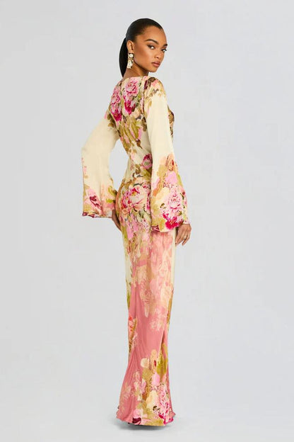 A model stands wearing a long-sleeved, floor-length column dress with a bateau neckline and dramatic flared bell sleeves. The cream-colored gown is adorned with a large-scale painterly floral print in shades of pink, rose, and olive green.