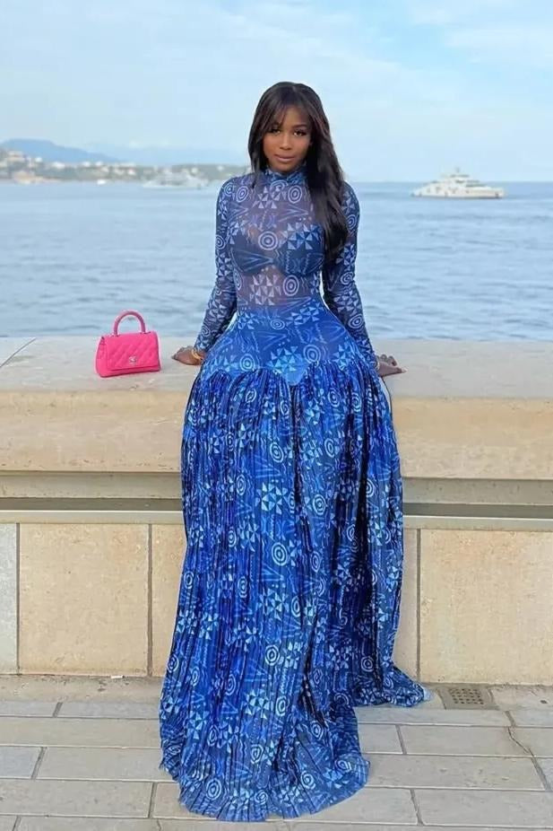 A woman sits on a stone ledge by the sea, wearing a long-sleeved, mock neck maxi dress in a blue and white geometric print. The dress features a sheer, fitted bodice and a voluminous, finely pleated skirt that pools around her.