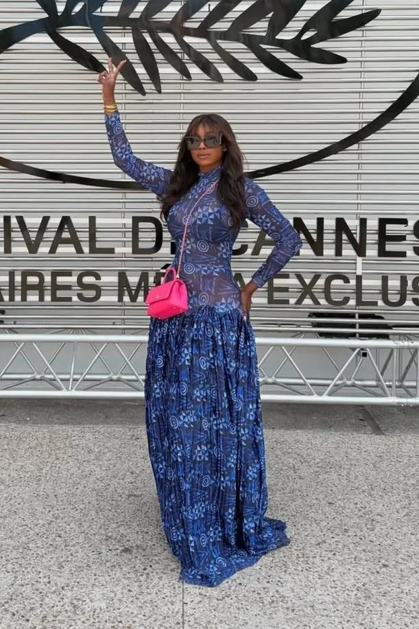 A woman sits on a stone ledge by the sea, wearing a long-sleeved, mock neck maxi dress in a blue and white geometric print. The dress features a sheer, fitted bodice and a voluminous, finely pleated skirt that pools around her.