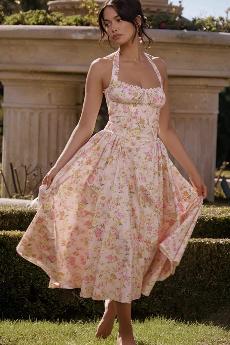 Pink - A woman with her hair in an updo smiles as she twirls in a sunlit garden, wearing a pink floral print halterneck midi dress with a corset-style ruched bodice and a full, flared skirt.