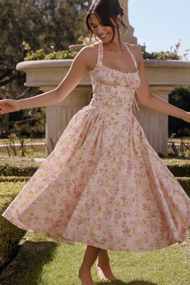 Pink - A woman with her hair in an updo smiles as she twirls in a sunlit garden, wearing a pink floral print halterneck midi dress with a corset-style ruched bodice and a full, flared skirt.