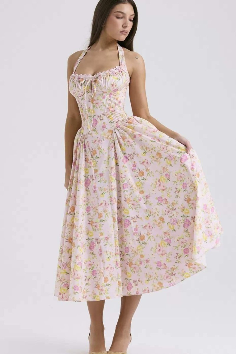 Pink - A woman with her hair in an updo smiles as she twirls in a sunlit garden, wearing a pink floral print halterneck midi dress with a corset-style ruched bodice and a full, flared skirt.