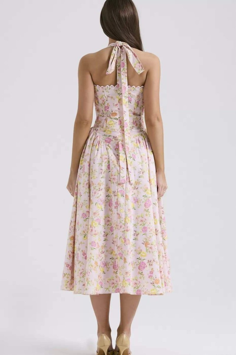 Pink - A woman with her hair in an updo smiles as she twirls in a sunlit garden, wearing a pink floral print halterneck midi dress with a corset-style ruched bodice and a full, flared skirt.