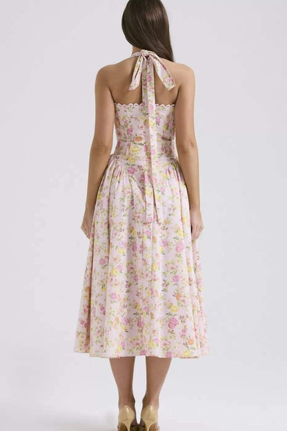 Pink - A woman with her hair in an updo smiles as she twirls in a sunlit garden, wearing a pink floral print halterneck midi dress with a corset-style ruched bodice and a full, flared skirt.